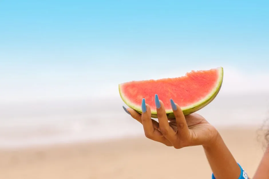 Person holder et stykke vandmelon med sommernegle 2026 i koraltone